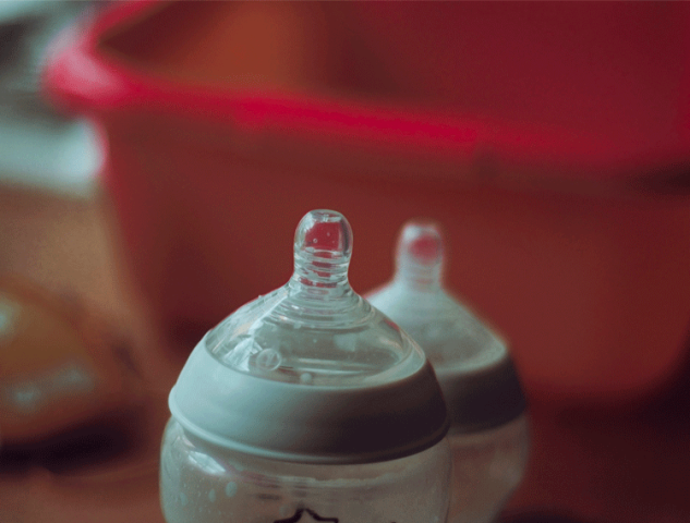 A close-up, out-of-focus shot of two baby bottles, with a red wash basin in the background, representing infant feeding routines at daycare.