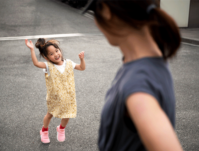 A young girl in a yellow dress smiles and dances playfully for an adult who is seen from behind.