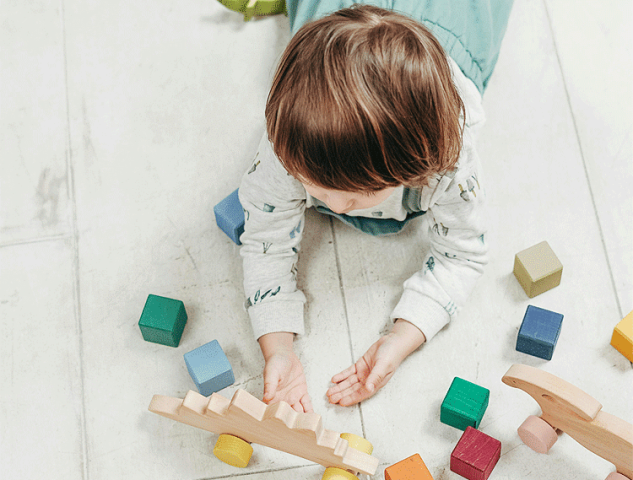 An overhead view of a young child playing with colorful wooden blocks and toys on a light-colored floor.