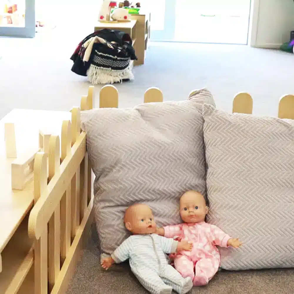 An early childhood education classroom with two baby dolls resting on gray chevron-patterned pillows inside a low, wooden fenced area.