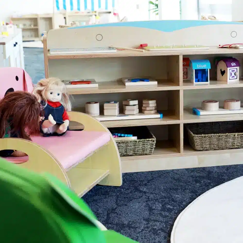 An early childhood education classroom with light wooden shelving, educational toys, a pink doll chair, and a doll sitting on the furniture.
