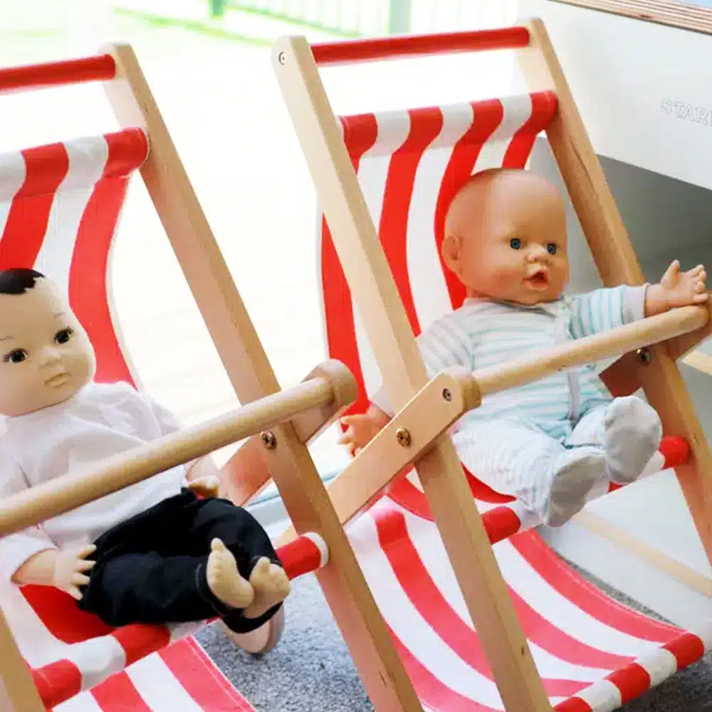 Two baby dolls sitting in miniature wooden deck chairs with red and white striped fabric near a window in an early childhood education classroom.