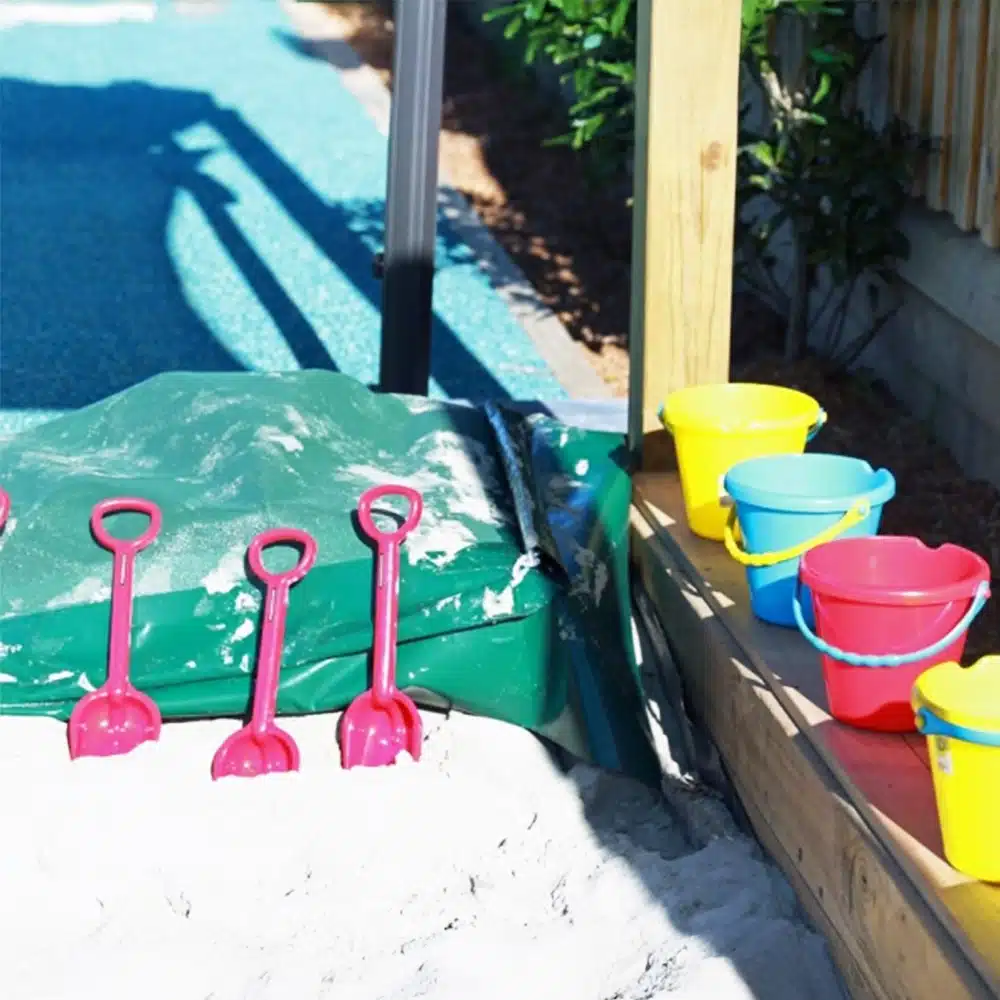 A sandbox area in a playground with white sand, pink plastic shovels, and brightly colored yellow, blue, and red buckets lined up on the wooden edge.