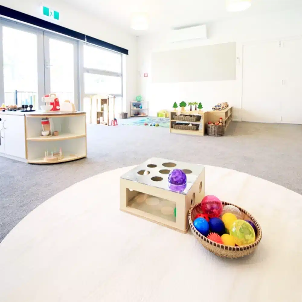 A bright, minimalist early childhood classroom interior with large windows, light colored furniture, and a central round play mat.